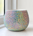 A round, planter pot covered in iridescent, crystal-embellished beads that reflect pastel rainbow colors, set on a white windowsill.