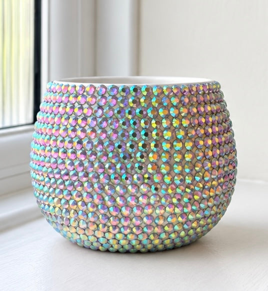 A round, planter pot covered in iridescent, crystal-embellished beads that reflect pastel rainbow colors, set on a white windowsill.