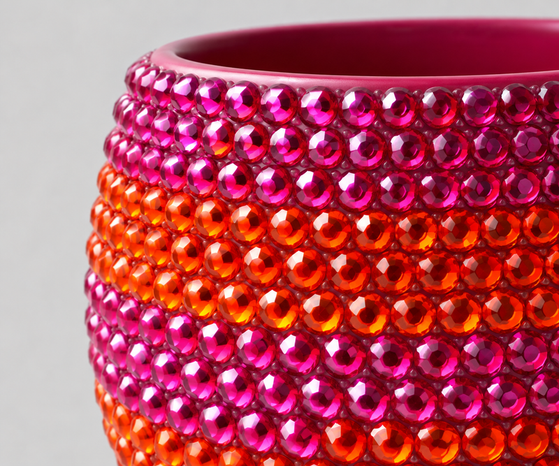 Close-up of a glossy ceramic plant pot covered in faceted crystals, showing a color gradient from magenta pink to bright orange against a light gray background.