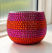 A round ceramic plant pot covered in rows of faceted crystals, featuring horizontal stripes of magenta pink and orange set on a windowsill.