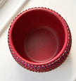 A top-down view of a small magenta ceramic plant pot decorated with sparkling fuchsia and orange crystals around its exterior, set against a clean white background.