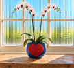 A blue ceramic pot with an orange heart design, holding a white orchid plant, set on a wooden windowsill with a bright window background.