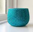 A round turquoise ceramic plant decorated with sparkling turquoise crystals shown sitting on a white windowsill with natural light coming from the left.