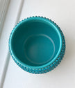 A turquoise blue ceramic plant pot with a textured, beaded outer rim, shown from a top-down perspective on a white paneled surface.