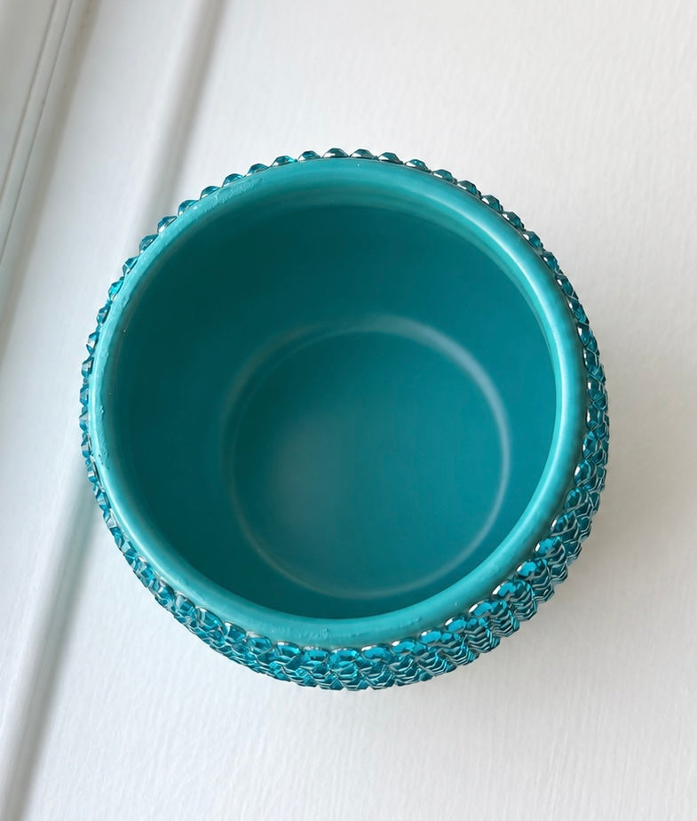 A turquoise blue ceramic plant pot with a textured, beaded outer rim, shown from a top-down perspective on a white paneled surface.