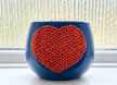A glossy blue planter pot with a large, textured crystal orange heart design on the front, set on a windowsill with a window blind background.