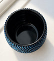 A small round black ceramic plant pot with a textured outer outer edge covered in metallic blue crystals, photographed from above on a light neutral surface.