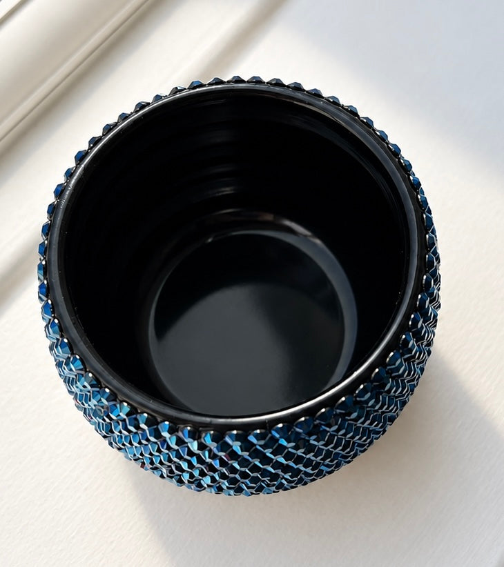 A small round black ceramic plant pot with a textured outer outer edge covered in metallic blue crystals, photographed from above on a light neutral surface.