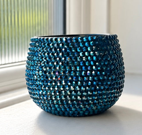 A small round decorative ceramic plant pot covered in shimmering blue and navy crystals, placed on a windowsill with natural light.