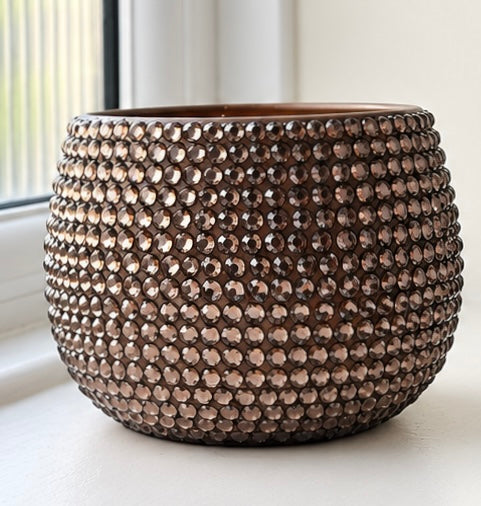 A round, dark brown planter covered in rows of small, faceted brown glass beads, set on a windowsill with natural light.