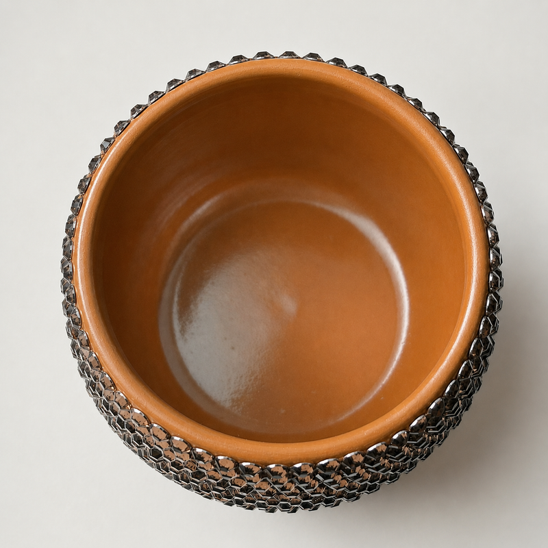 A top-down view of a round ceramic plant pot with a glossy light brown interior and a dark metallic chainmail-like textured exterior, set against a plain light gray background.