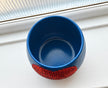 A blue ceramic plant pot with a crystal orange heart in the middle  photographed on a white windowsill with a window pane visible in the background.