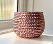 A round ceramic plant pot covered in horizontal rows of pink and gold crystals, sitting on a windowsill with natural light.