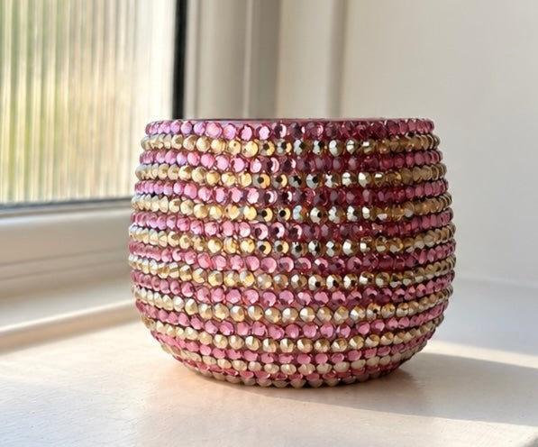 A round ceramic plant pot covered in horizontal rows of pink and gold crystals, sitting on a windowsill with natural light.