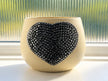 A cream-colored ceramic planter with a large, textured grey heart-shaped embellishment on the front, set on a windowsill with a ribbed glass window pane in the background.