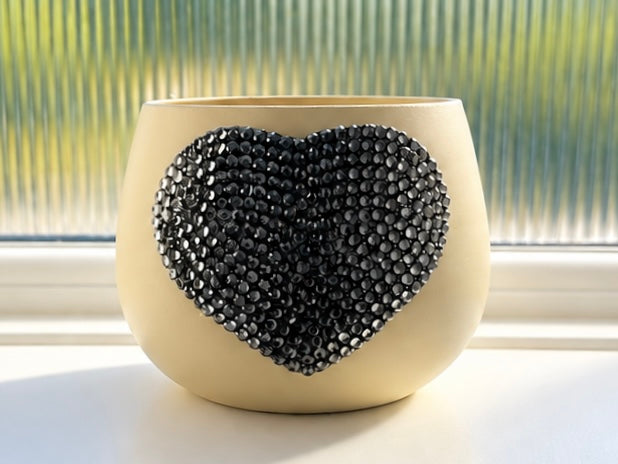 A cream-colored ceramic planter with a large, textured grey heart-shaped embellishment on the front, set on a windowsill with a ribbed glass window pane in the background.