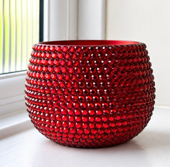A round, glossy cerramic planter  covered in rows of faceted red crystals, set on a white windowsill with a window and white wall in the background.