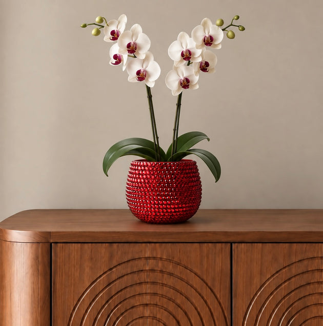 A crimson red ceramic plant pot decorated with sparkling ruby red crystals containing a white orchid. Pot is shown sitting on a brown sideboard. 