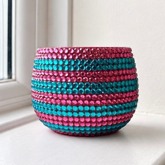 A round ceramic plant pot covered in horizontal stripes of pink and teal crystals, sitting on a white windowsill.