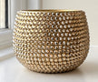 A round, gold-toned decorative planter  covered in tightly packed faceted beads, photographed on a white windowsill with a window and soft natural light in the background.