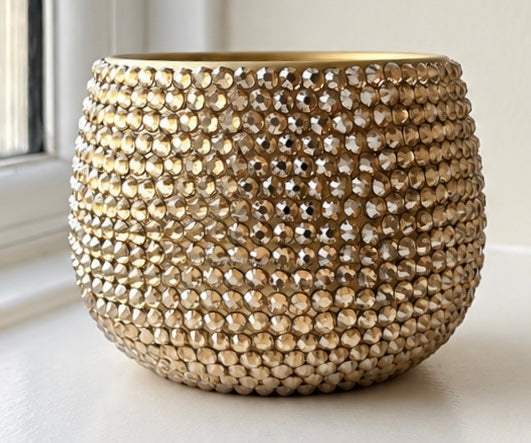 A round, gold-toned decorative planter  covered in tightly packed faceted beads, photographed on a white windowsill with a window and soft natural light in the background.