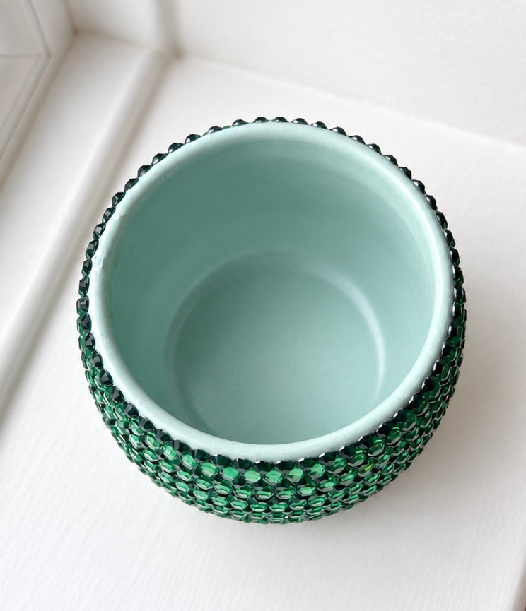 A small round ceramic plant pot with a glossy light aqua interior and a textured outer rim covered in dark green faceted crystals, shown from a top-down angle on a white surface.