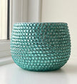 A round jade planter covered in small faceted crystals, sitting on a white windowsill with a window and wall visible in the background.