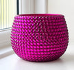 A round, ceramic planter in a vibrant magenta pink, covered in a dense pattern of small, raised circular facets. It is photographed sitting on a white windowsill with a window and white wall visible in the background.
