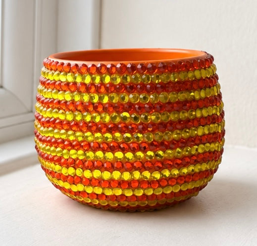 A small round ceramic planter pot decorated with horizontal stripes of faceted orange and yellow crystals, set on a white surface near a window.