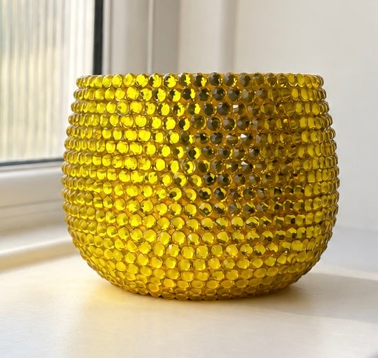 A round, textured yellow planter decorated with sparkling yellow crystals  sitting on a windowsill with natural light.
