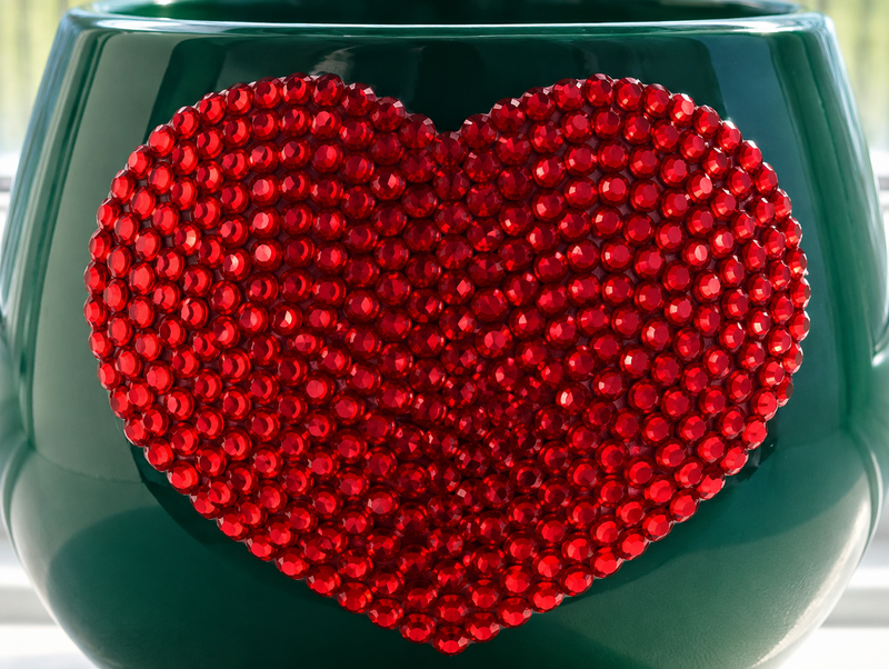 A close-up of a dark green glossy ceramic plant pot decorated with a large red heart-shaped embellishment covered in small red crystals
