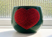 A dark green ceramic planter pot with a large red heart-shaped crystal in the center, placed on a windowsill with a frosted window in the background.