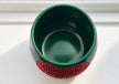 A dark green ceramic plant pot with a textured red crystal heart in the middle photographed from a top-down angle on a white surface.
