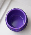 A purple ceramic plant pot with a textured, beaded outer rim, photographed from a top-down angle on a white surface.