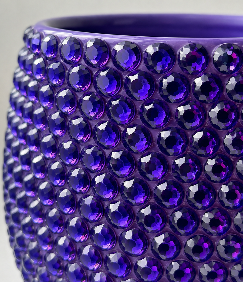 Close-up of a purple plant pot covered in faceted round purple crystals, showing the glossy textured surface and curved rim against a light neutral background.