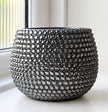A round, dark grey ceramic pot covered in a dense, textured pattern of faceted grey crystals, set on a white windowsill with a window and white wall visible in the background.