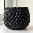 A black textured ceramic planter with a raised, faceted bead-like pattern, sitting on a white windowsill with natural light.