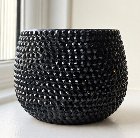 A black textured ceramic planter with a raised, faceted bead-like pattern, sitting on a white windowsill with natural light.