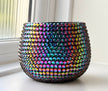 A round, wide-mouthed ceramic plant pot covered in black iridescent, rainbow-colored crystals, sitting on a windowsill.