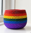 A round ceramic plant pot decorated with horizontal rainbow stripes of red, orange, yellow, green, blue, and purple crystals, set on a white windowsill.
