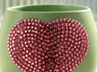 A close-up of a light sage green planter decorated with a large, textured heart-shaped embellishment covered in sparkling rose pink crystals.