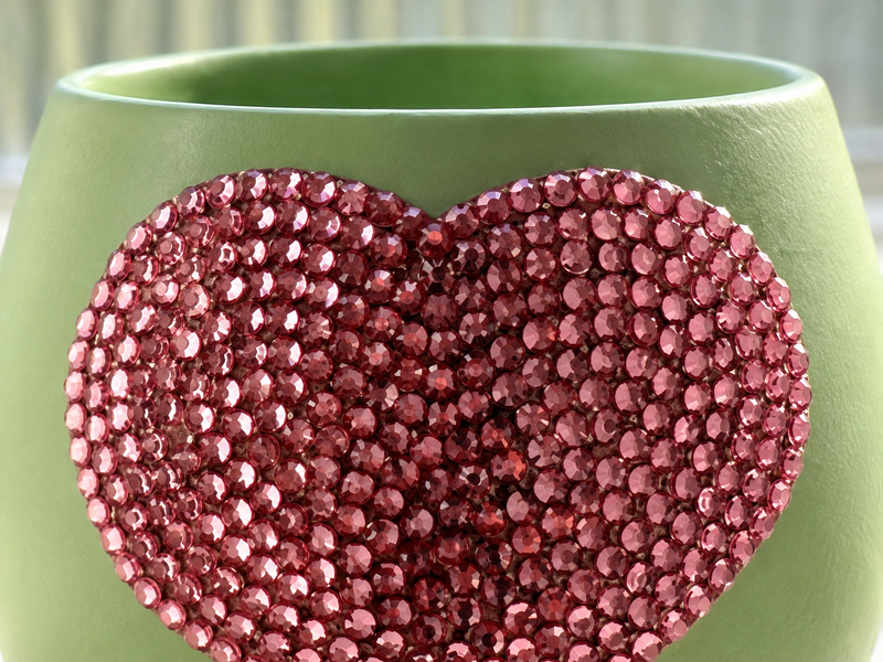 A close-up of a light sage green planter decorated with a large, textured heart-shaped embellishment covered in sparkling rose pink crystals.