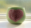 A light sage green ceramic planter with a large, textured, rose-gold crystal-embellished heart on the front, set on a windowsill with a ribbed glass window pane in the background.