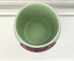 A top-down view of a light sage green ceramic plant pot with a textured, pinkish crystal-embellished heart, set against a white background.