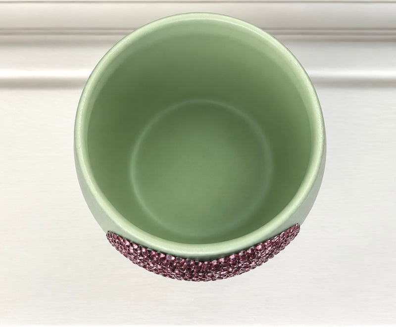 A top-down view of a light sage green ceramic plant pot with a textured, pinkish crystal-embellished heart, set against a white background.