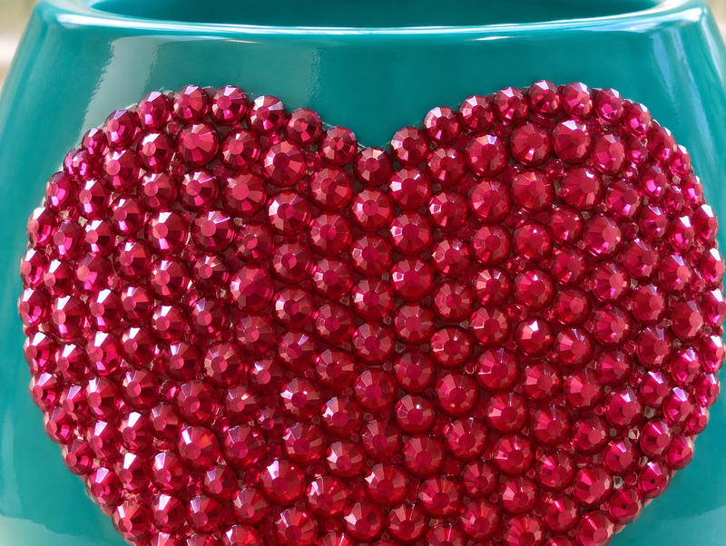 A close-up of a glossy teal ceramic plant pot decorated with a large heart-shaped arrangement of hot pink crystals