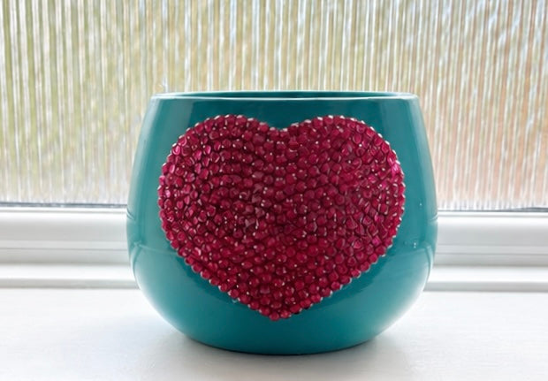 A teal ceramic planter pot with a large, textured, fuchsia pink crystal heart design on the front, set on a windowsill with a window blind background.