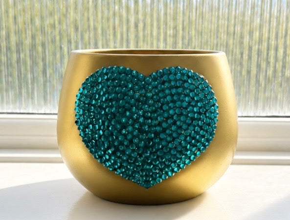 A gold-colored ceramic plant pot with a large, textured teal heart-shaped embellishment on the front, set on a windowsill with natural light.