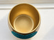 A gold-colored plant pot with a textured, glittery teal blue heart in the centre photographed from a top-down angle on a white surface.