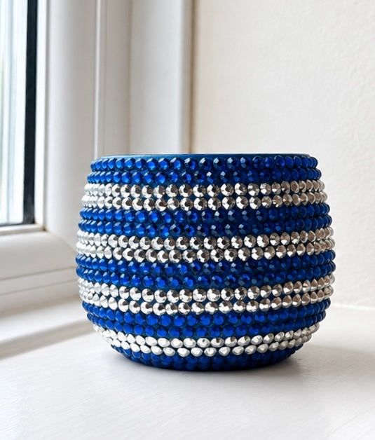 A small round decorative planter covered in horizontal stripes of faceted blue and silver crystals, sitting on a windowsill.